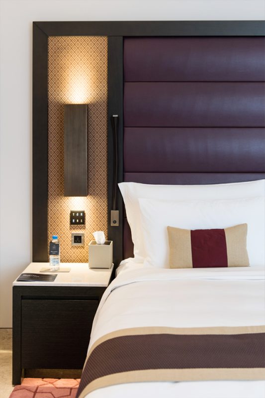 Hotel Bedroom Design - Padded velvet purple headboard & dark wood pedestal below lighting feature