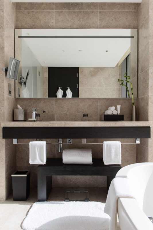 Bathroom Interior Design - Textured, tiled walls & countertop with dark, solid wood furniture next to frosted glass door